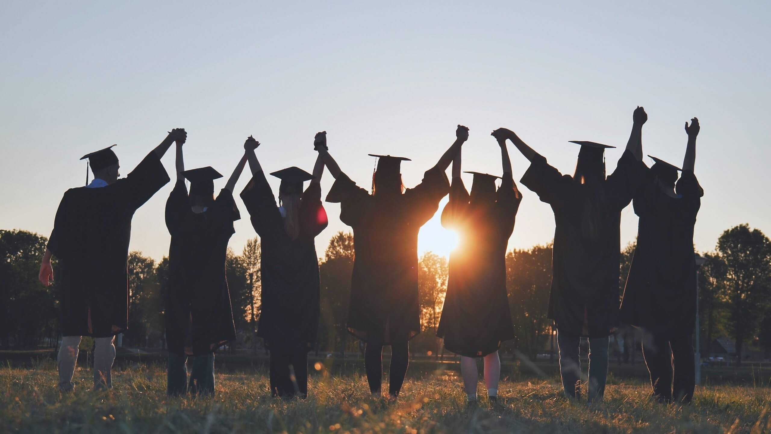 College,Graduates,In,Robes,Holding,Hands,At,Sunset.